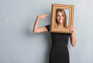 Beautiful young woman over grunge grey wall holding vintage frame very happy pointing with hand and...