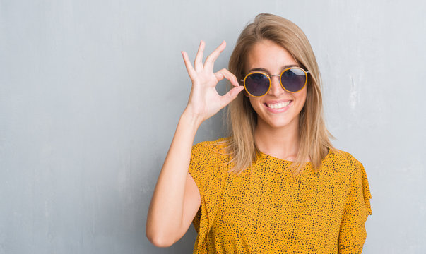 Beautiful Young Woman Standing Over Grunge Grey Wall Wearing Retro Sunglasses Doing Ok Sign With Fingers, Excellent Symbol