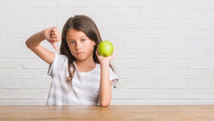 Young hispanic kid sitting on the table eating fresh green apple with angry face, negative sign showing dislike with thumbs down, rejection concept