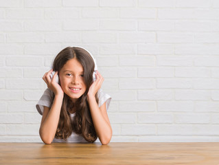 Young hispanic kid sitting on the table wearing headphones with a happy face standing and smiling with a confident smile showing teeth