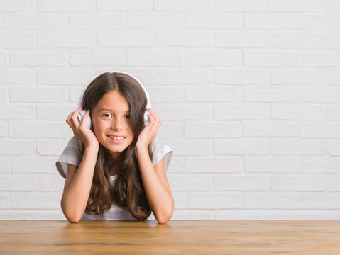Young hispanic kid sitting on the table wearing headphones with a happy face standing and smiling with a confident smile showing teeth