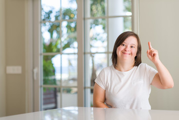 Fototapeta premium Down syndrome woman at home pointing finger up with successful idea. Exited and happy. Number one.