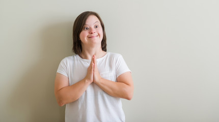Down syndrome woman standing over wall begging and praying with hands together with hope expression...