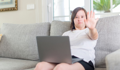 Obraz na płótnie Canvas Down syndrome woman at home using computer laptop with open hand doing stop sign with serious and confident expression, defense gesture
