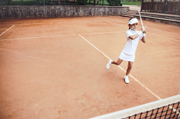 Obraz premium Child learning to play tennis in the sport club. Full length shot of a little girl tennis player on clay court. Sporty child girl hits the ball with a racket. Tennis training for young kid.