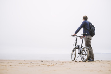 A young man with a backpack standing with the bike on the beach looking at the sea.
