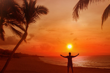 Silhouette of a man with his arms outstretched. Behind him is a beautiful sunset over the sea and the beach with palms.