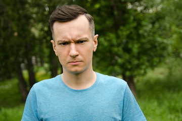 Young unhappy guy looks in a camera a green grass and trees background.