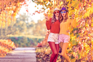 Fall Fashion. Two Young Amazing Woman in Colorful Park with Kiss Face and peace sign. Lovable Girl Friends Fooling Around Enjoy Nature. Urban Outdoor. Model in Stylish Trendy Autumn Outfit