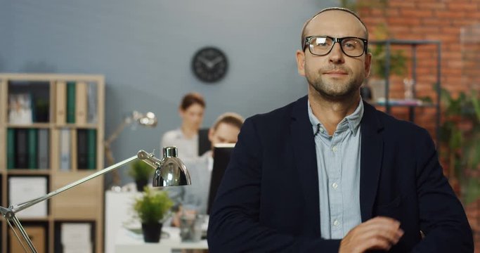 Portrait shot of the Caucasian handsome businessman ib glasses and official style standing in the office room, leaning on the table and turning head to the camera.