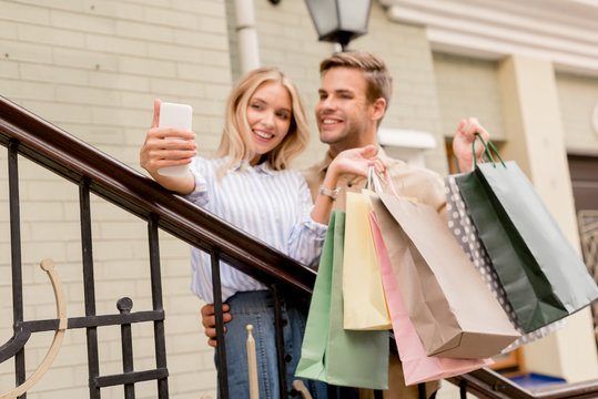 smiling young couple with paper bags taking selfie on smartphone near store at city street