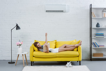 relaxed young woman reading book on sofa and pointing at air conditioner with remote control