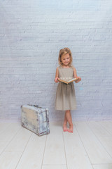 the girl, baby in dress on white brick wall background with suitcase, with a book