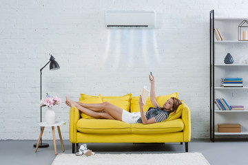 concentrated young woman reading book on couch and pointing at air conditioner hanging on wall with...