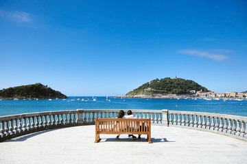 Fototapeta premium Zakochana para siedzi na drewnianej ławce z widokiem na morze. Letni słoneczny dzień z błękitnym niebem. San Sebastian lub Donostia, Kraj Basków, Hiszpania