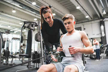 Young man in gym with his personal trainer working out on cable weight machine. - obrazy, fototapety, plakaty