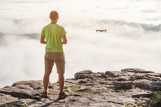 Young Man navigating a flying drone at fog and clouds, standing on top of the mountains at sunset