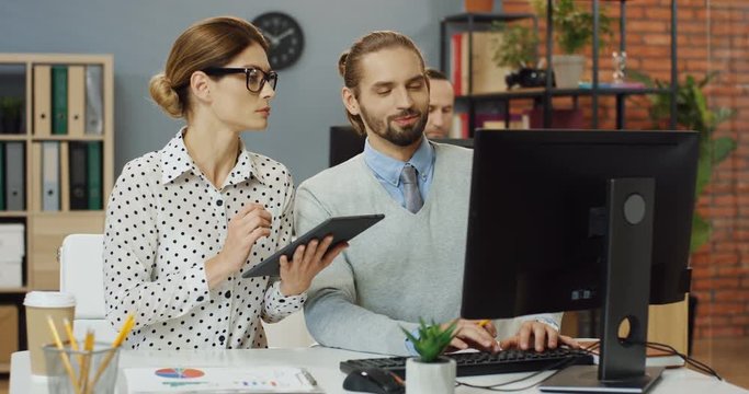 handsome man sitting in the office and working on the laptop computer while seniour female co-worker taping on the tablet and helping him.