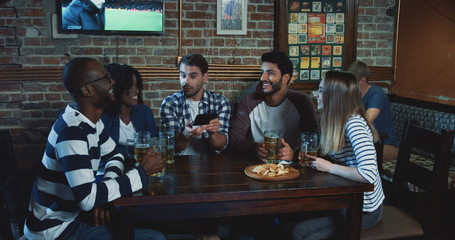 Team of mixed races male and female friends sitting in the pub and watching sport game on the smartphone, celebrating victory loudly.
