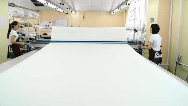 Two female factory workers unrolling white cotton fabric and cutting it with automatic circular blade of textile machine
