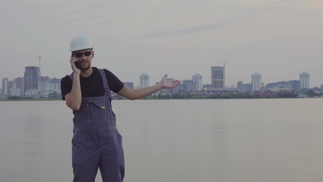 4K shot of handsome caucasian worker in white helmet talking on mobile phone near the beautiful evening city and construction site. construction success