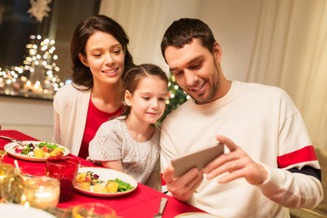 Obraz na płótnie Canvas holidays, family and technology concept - happy mother, father and little daughter with smartphone having christmas dinner at home