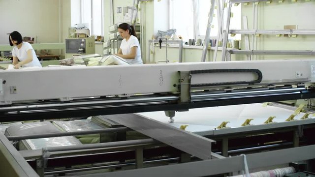 Tracking shot of automated machine quilting white fabric while Asian female workers packing blankets in textile factory