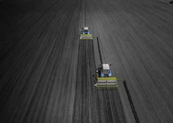 Aerial view of two blue tractors plows the earth in field on a summer day against a black earth...