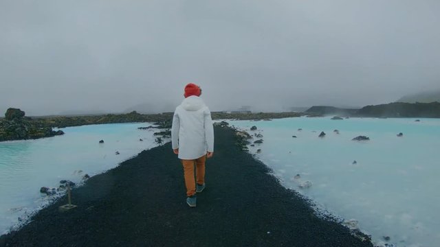 Hipster millennial man in white raincoat and bright orange beanie hat, explores epic terrain in iceland volcanic hot springs, amazing travel destination for adventure seekers, nomad lifestyle
