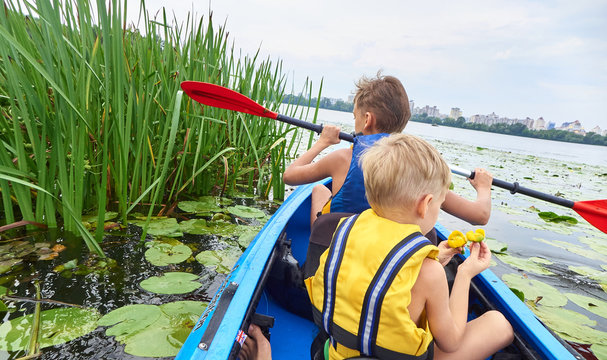 Children boys swim on a plastic kayak boat on the lake with water lilies.