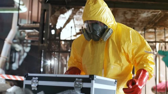 Man in gas mask and new yellow protective suit opening black briefcase. Guy wearing red rubber gloves looking for something. Indoors.