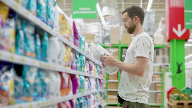 Casual adult man with beard holding big bag of pet food in store, man choosing food for domestic animal. Customer making decision in supermarket.