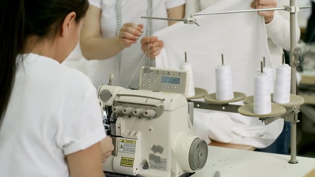 Female worker sitting at workplace and using overlock sewing machine while two seamstresses discussing fabric
