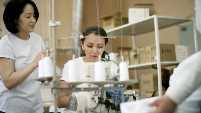 Tracking shot of experienced Asian seamstress explaining young female colleague how to use overlock sewing machine, smiling and giving high five while working together in textile factory