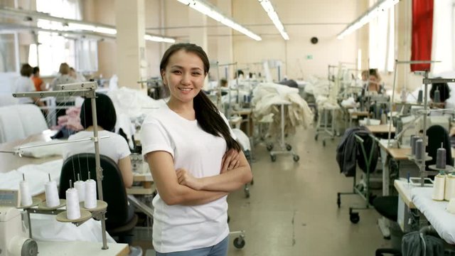 Beautiful young Asian woman standing with arms crossed, looking at camera and smiling while working in sewing factory