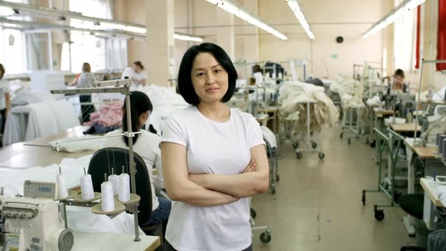 Zoom in of middle-aged female Asian worker posing at camera in sewing factory during workday