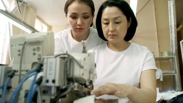 Professional Asian seamstress teaching female colleague how to use sewing machine while working together in textile factory