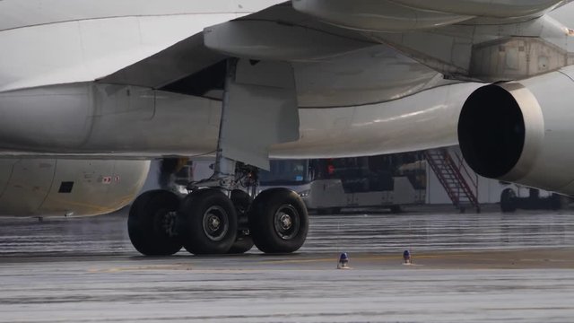 Landing gear and engine of jetliner taxiing on wet airport ramp. Sunlight spots on fuselage
