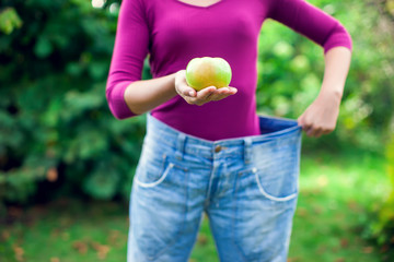 Young woman wearing big loose jeans with apple in hand - weight loss concept