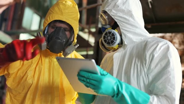 Two Caucasian men wearing yellow and white hazmat suits working together in old building. Modern scientists using digital tablet. Contamination. Indoors.