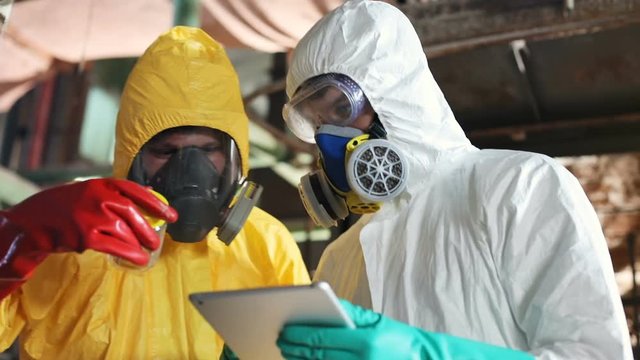 Two Caucasian students in colored hazmat outfits doing tests. Young people using digital tablet. Guys in gas masks looking directly at camera. Indoors.