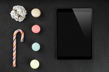 Digital tablet with sweet macarons, lollipop and biscuit on stone background. Copy space, flat lay