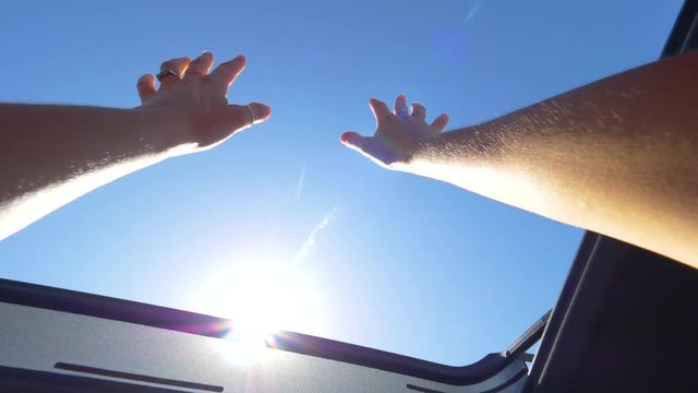 SLOW MOTION, CLOSE UP, POV, LENS FLARE: Cheerful woman with rings outstretching her arms through the sunroof of a car during her summer road trip. Carefree young female tourist enjoying a car cruise.