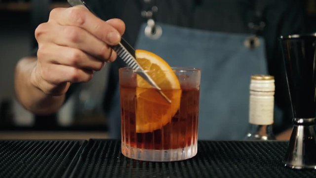 Close up shot of bartender hands preparing negroni cocktail with grapefruit. He is putting grapefruit skin into the cocktail glass on counter.
