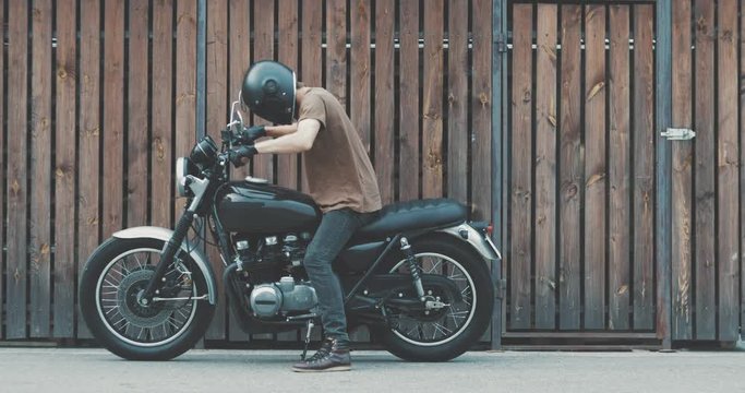 Handsome rider guy with beard and mustache parked classic style motorcycle near wooden wall of garage. Urban lifestyle 4k video