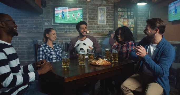 Cheerful multiethnical friends playing a ball while sitting at the table in the pub during sport game evening.