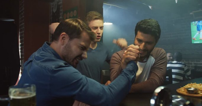 Two young attractive man having an arm wrestling match while sitting at the bar during sport game on TV.