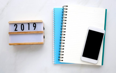 2019 on wood box, blank notebook paper and smart phone with blank screen on white marble table background, 2019 new year mock up, template with copy space for text, top view
