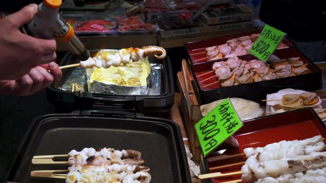 a vendor uses a gas torch to finish cooking octopus skewers at nishiki market in kyoto , japan