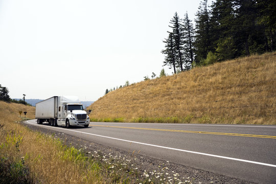Big rig white semi truck with trailer running on spectacular winding road with yellow grass hills